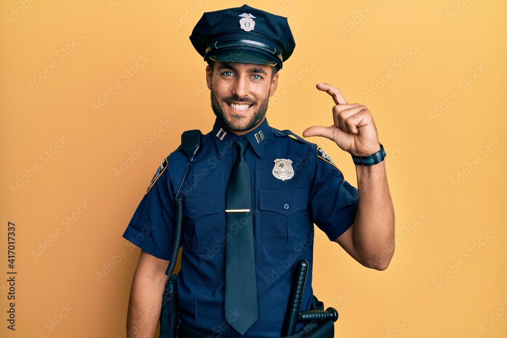 Handsome hispanic man wearing police uniform smiling and confident gesturing with hand doing small size sign with fingers looking and the camera. measure concept. Handsome hispanic man wearing police uniform smiling and confident gesturing with hand doing small size sign with fingers looking and the camera. measure concept.