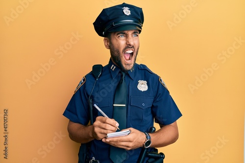 Fototapeta Handsome hispanic man wearing police uniform writing traffic fine angry and mad screaming frustrated and furious, shouting with anger