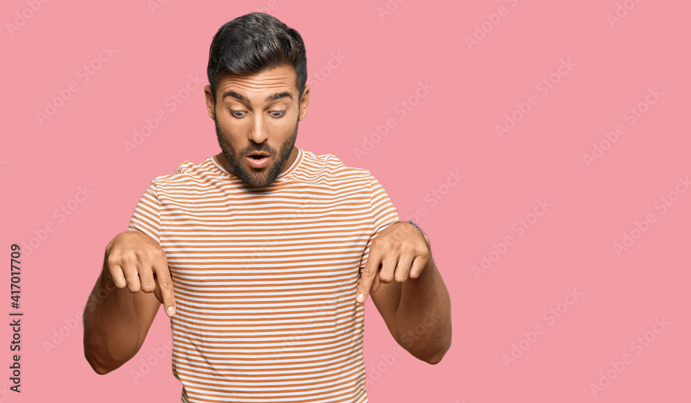 Handsome hispanic man wearing casual clothes pointing down with fingers showing advertisement, surprised face and open mouth Handsome hispanic man wearing casual clothes pointing down with fingers showing advertisement, surprised face and open mouth