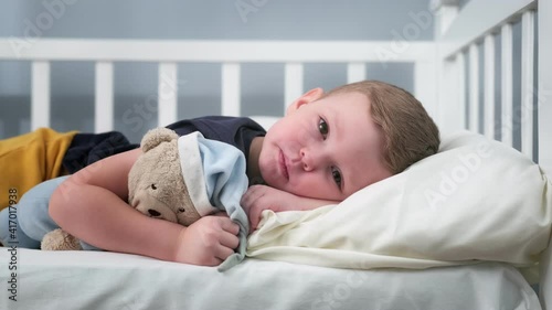 Small caucasian child boy depressed alone hugs teddy bear with frustrated and frightened look looking ahead of leda on bed. Concept of loneliness, fear, and domestic violence against children. photo
