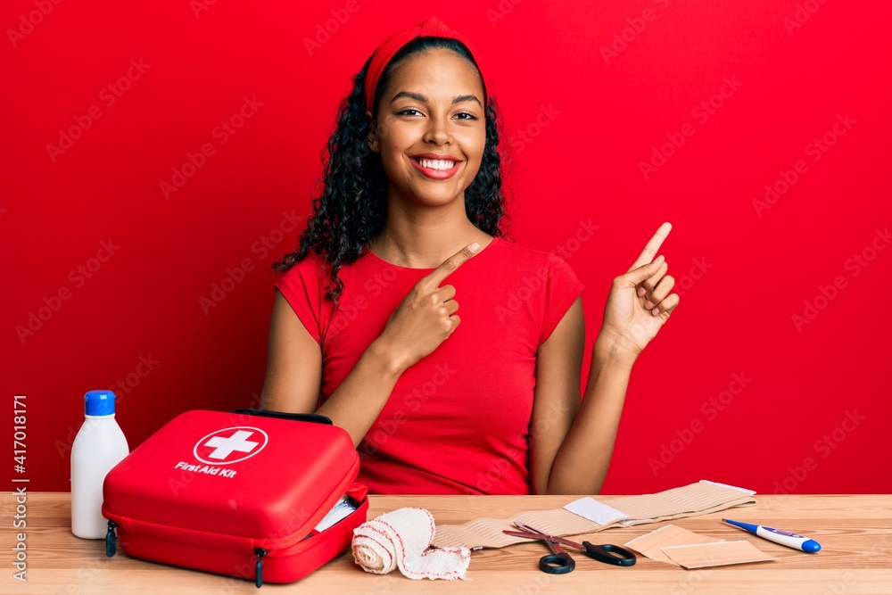 Young african american girl sitting on the table with first aid kit making cure smiling and looking at the camera pointing with two hands and fingers to the side. Young african american girl sitting on the table with first aid kit making cure smiling and looking at the camera pointing with two hands and fingers to the side.