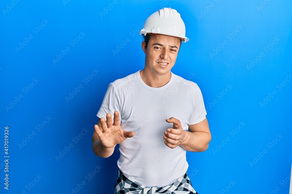Handsome young man wearing builder uniform and hardhat disgusted expression, displeased and fearful doing disgust face because aversion reaction. with hands raised