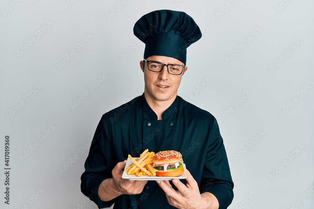 Handsome young man chef holding burger with fries in shock face, looking skeptical and sarcastic, surprised with open mouth Handsome young man chef holding burger with fries in shock face, looking skeptical and sarcastic, surprised with open mouth