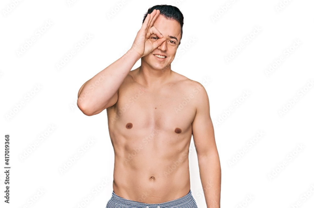 Fototapeta premium Handsome young man wearing swimwear shirtless doing ok gesture with hand smiling, eye looking through fingers with happy face.