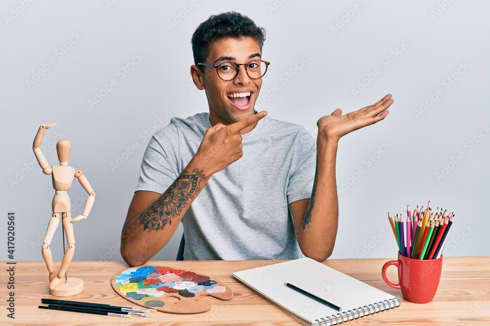 Young handsome african american man painter sitting palette and art manikin amazed and smiling to the camera while presenting with hand and pointing with finger. Young handsome african american man painter sitting palette and art manikin amazed and smiling to the camera while presenting with hand and pointing with finger.