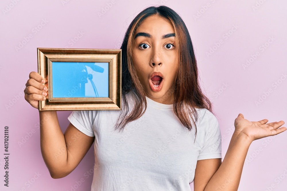 Young latin woman holding empty frame scared and amazed with open mouth for surprise, disbelief face Young latin woman holding empty frame scared and amazed with open mouth for surprise, disbelief face