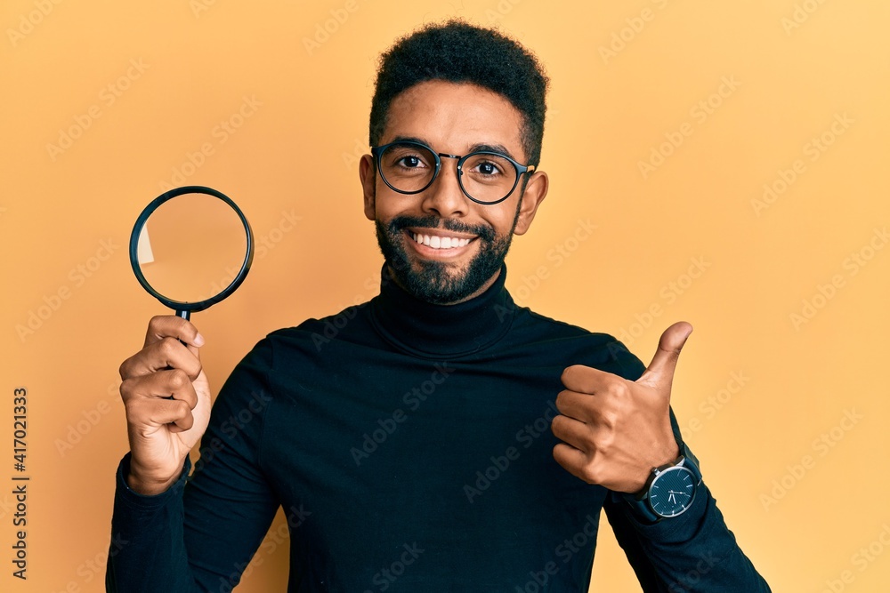 Handsome hispanic man with beard holding magnifying glass smiling happy and positive, thumb up doing excellent and approval sign Handsome hispanic man with beard holding magnifying glass smiling happy and positive, thumb up doing excellent and approval sign