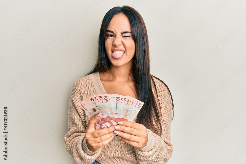 Young beautiful hispanic girl holding 10 colombian pesos banknotes sticking tongue out happy with funny expression. Young beautiful hispanic girl holding 10 colombian pesos banknotes sticking tongue out happy with funny expression.