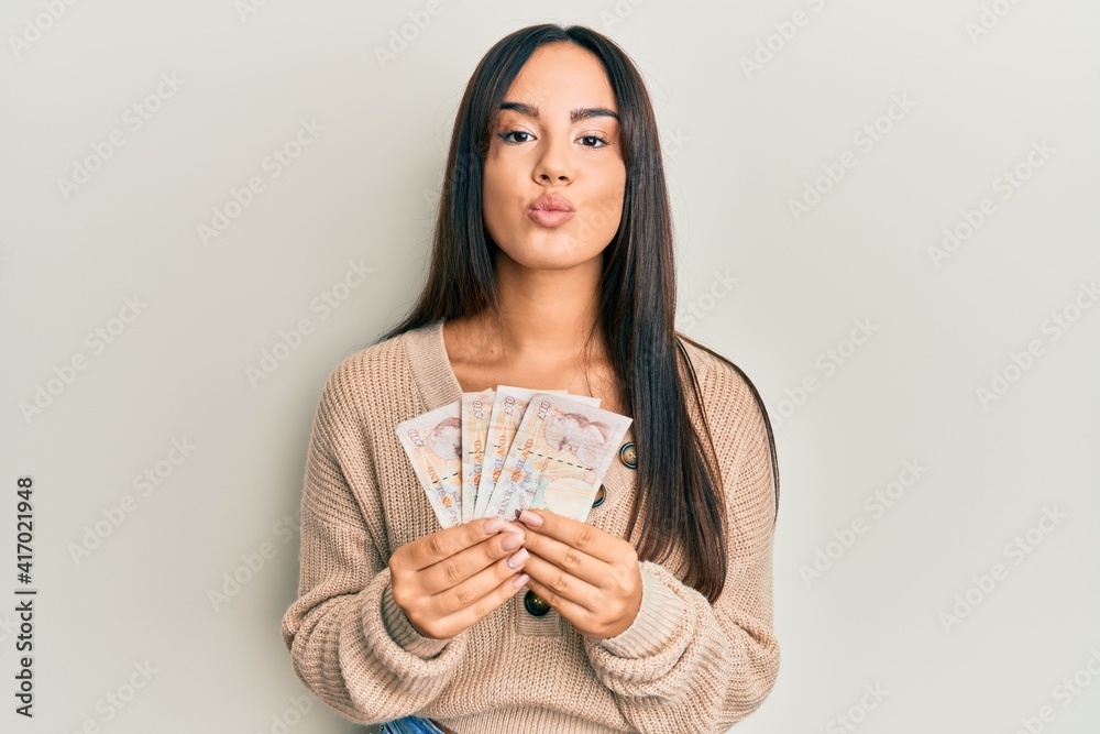Young beautiful hispanic girl holding 10 united kingdom pounds banknotes looking at the camera blowing a kiss being lovely and sexy. love expression. Young beautiful hispanic girl holding 10 united kingdom pounds banknotes looking at the camera blowing a kiss being lovely and sexy. love expression.