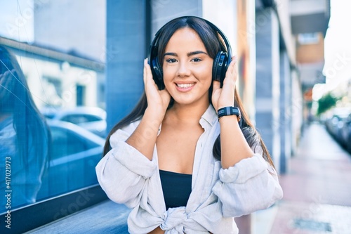 Young hispanic girl smiling happy listening to music using headphones at the city.