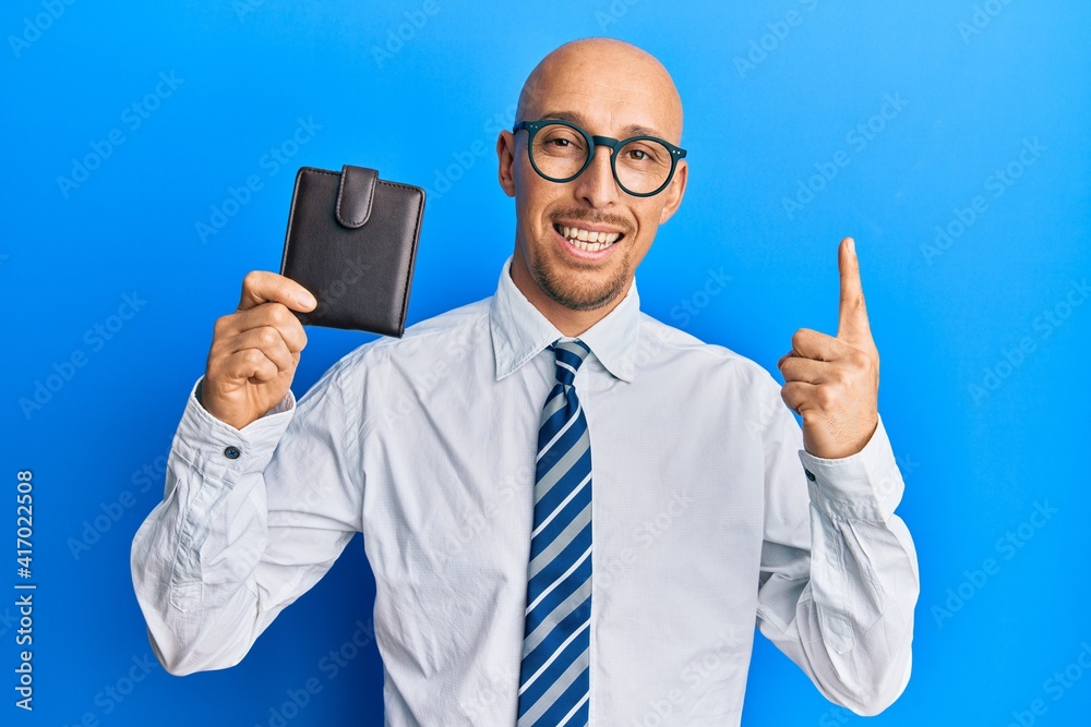 Bald man with beard holding leather wallet smiling with an idea or question pointing finger with happy face, number one Bald man with beard holding leather wallet smiling with an idea or question pointing finger with happy face, number one