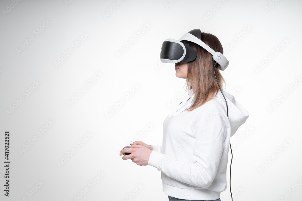 Teenage girl with fair hair is playing game with a gamepad and VR glasses in a white studio, glasses covers almost all her face, side view, waist high Teenage girl with fair hair is playing game with a gamepad and VR glasses in a white studio, glasses covers almost all her face, side view, waist high
