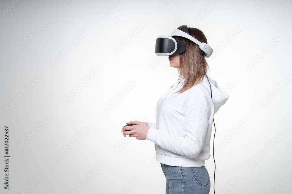 Teenage girl with fair hair is playing game with a gamepad and VR glasses in a white studio, glasses covers almost all her face, side view, waist high Teenage girl with fair hair is playing game with a gamepad and VR glasses in a white studio, glasses covers almost all her face, side view, waist high