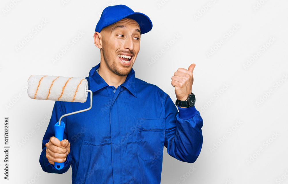 Bald man with beard holding roller painter pointing thumb up to the side smiling happy with open mouth