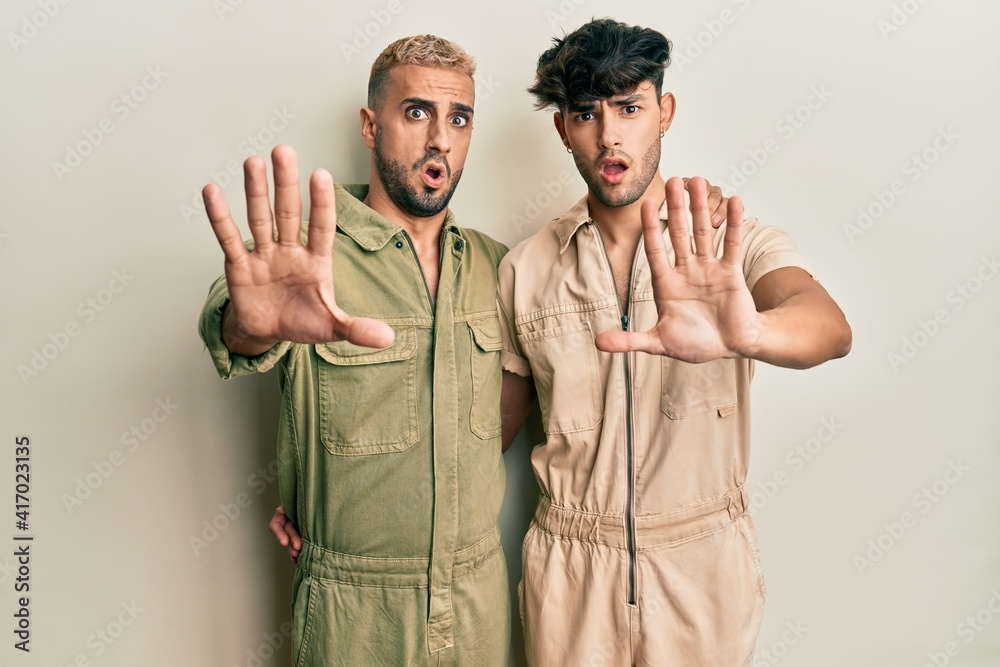 Homosexual gay couple standing together wearing casual jumpsuit doing stop gesture with hands palms, angry and frustration expression