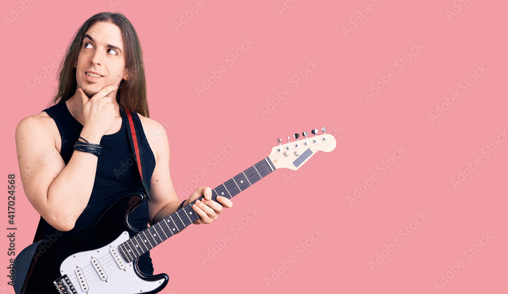 Young adult man with long hair playing electric guitar serious face thinking about question with hand on chin, thoughtful about confusing idea Young adult man with long hair playing electric guitar serious face thinking about question with hand on chin, thoughtful about confusing idea