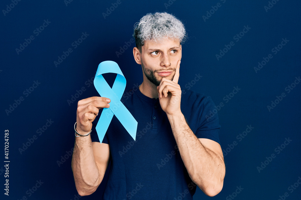 Obraz premium Young hispanic man with modern dyed hair holding pink cancer ribbon serious face thinking about question with hand on chin, thoughtful about confusing idea