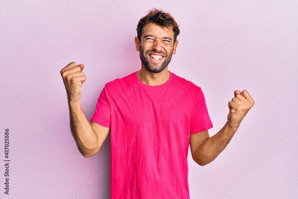 Handsome man with beard wearing casual pink tshirt over pink background screaming proud, celebrating victory and success very excited with raised arms