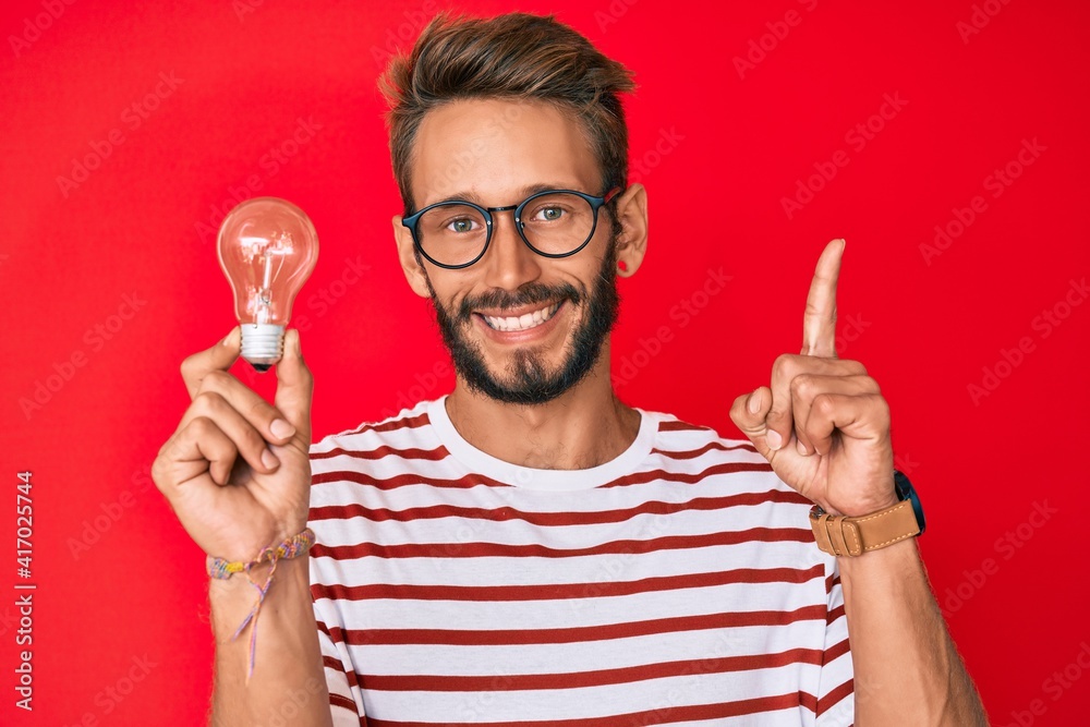 Handsome caucasian man with beard holding lightbulb for inspiration and idea surprised with an idea or question pointing finger with happy face, number one Handsome caucasian man with beard holding lightbulb for inspiration and idea surprised with an idea or question pointing finger with happy face, number one
