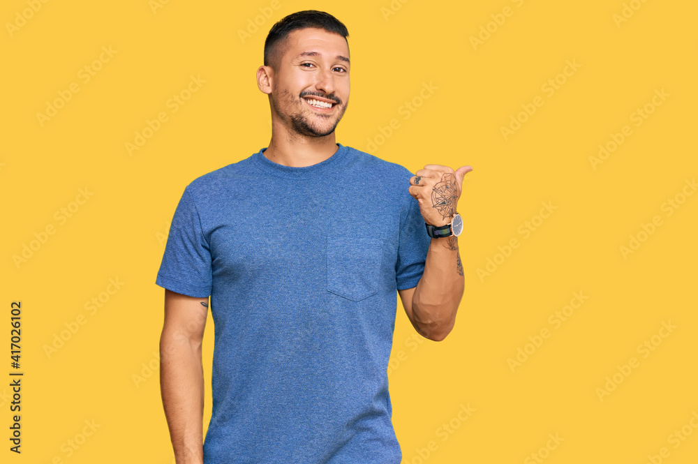 Handsome man with tattoos wearing casual clothes smiling with happy face looking and pointing to the side with thumb up. Handsome man with tattoos wearing casual clothes smiling with happy face looking and pointing to the side with thumb up.