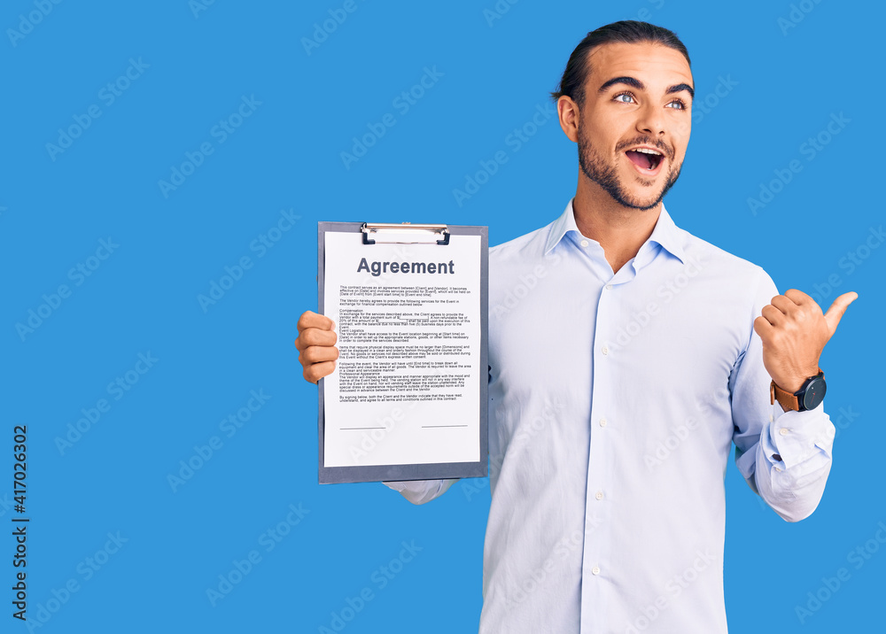 Young handsome man holding clipboard with agreement document pointing thumb up to the side smiling happy with open mouth