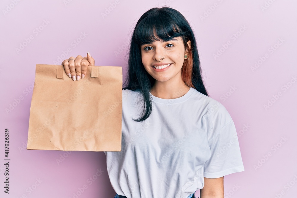 Young hispanic girl holding take away paper bag looking positive and happy standing and smiling with a confident smile showing teeth Young hispanic girl holding take away paper bag looking positive and happy standing and smiling with a confident smile showing teeth