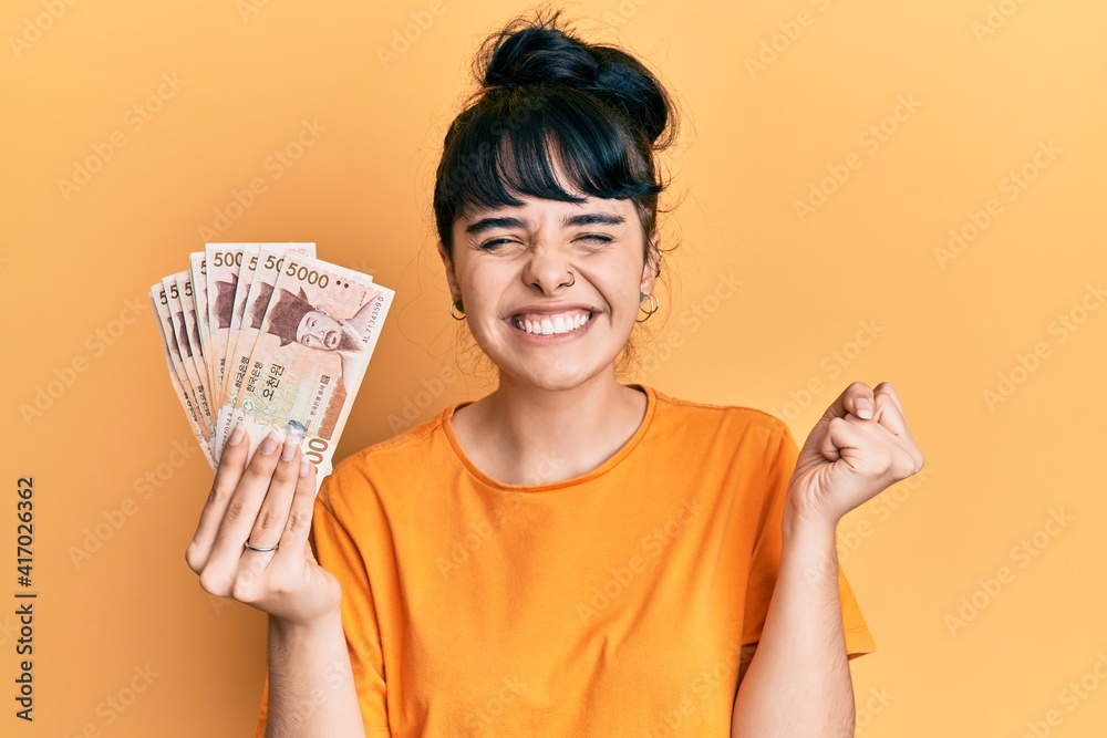 Young hispanic girl holding 5000 south korean won banknotes screaming proud, celebrating victory and success very excited with raised arm Young hispanic girl holding 5000 south korean won banknotes screaming proud, celebrating victory and success very excited with raised arm