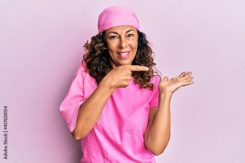 Middle age hispanic woman wearing breast cancer support pink scarf amazed and smiling to the camera while presenting with hand and pointing with finger. Middle age hispanic woman wearing breast cancer support pink scarf amazed and smiling to the camera while presenting with hand and pointing with finger.