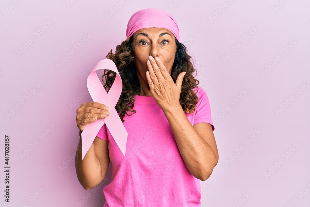 Middle age hispanic woman holding pink cancer ribbon covering mouth with hand, shocked and afraid for mistake. surprised expression Middle age hispanic woman holding pink cancer ribbon covering mouth with hand, shocked and afraid for mistake. surprised expression