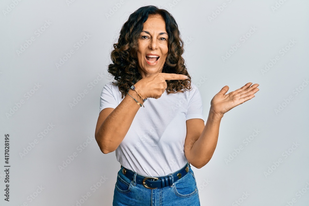 Middle age hispanic woman wearing casual white t shirt amazed and smiling to the camera while presenting with hand and pointing with finger. Middle age hispanic woman wearing casual white t shirt amazed and smiling to the camera while presenting with hand and pointing with finger.