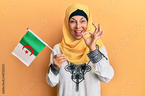 Middle age hispanic woman wearing hijab holding algeria flag doing ok sign with fingers, smiling friendly gesturing excellent symbol