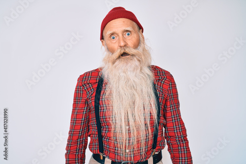 Old senior man with grey hair and long beard wearing hipster look with wool cap in shock face, looking skeptical and sarcastic, surprised with open mouth