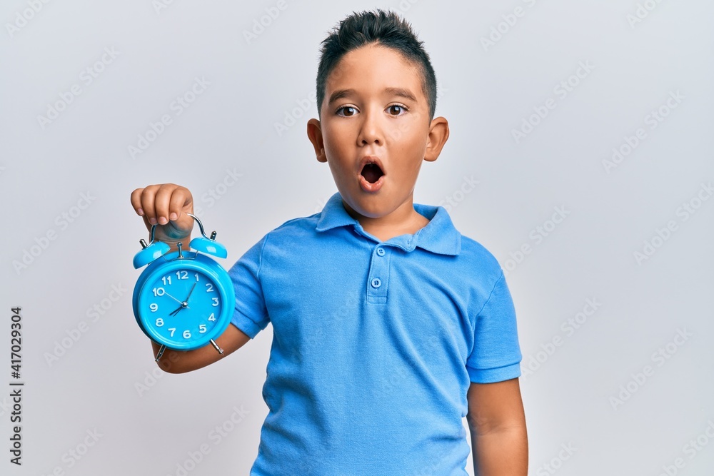 Little boy hispanic kid holding alarm clock scared and amazed with open mouth for surprise, disbelief face Little boy hispanic kid holding alarm clock scared and amazed with open mouth for surprise, disbelief face