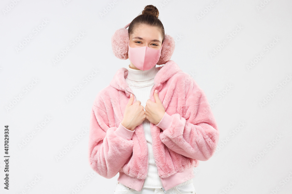 Young woman wearing fur coat, earmuffs and pink mask to protect from covid virus, isolated on gray background