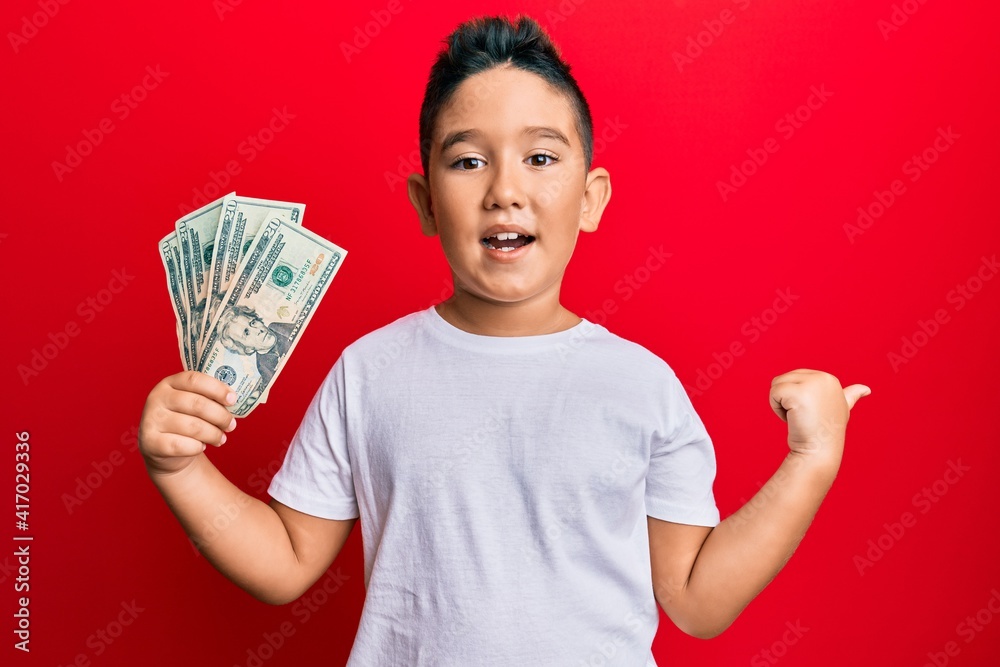Little boy hispanic kid holding 20 dollars banknotes pointing thumb up to the side smiling happy with open mouth Little boy hispanic kid holding 20 dollars banknotes pointing thumb up to the side smiling happy with open mouth