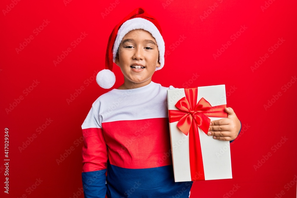 Little boy hispanic kid wearing christmas hat and holding a gift looking positive and happy standing and smiling with a confident smile showing teeth Little boy hispanic kid wearing christmas hat and holding a gift looking positive and happy standing and smiling with a confident smile showing teeth