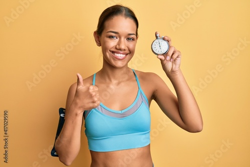 Beautiful hispanic woman wearing sportswear holding countdown clock smiling happy and positive  thumb up doing excellent and approval sign