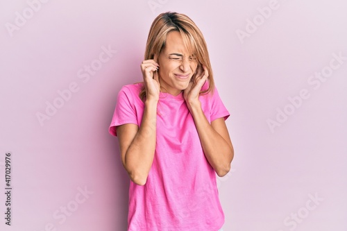 Beautiful blonde woman wearing casual pink tshirt covering ears with fingers with annoyed expression for the noise of loud music. deaf concept.