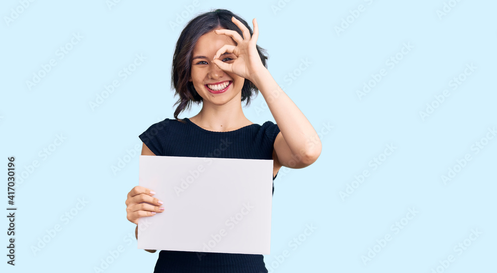 Young beautiful girl holding blank empty banner smiling happy doing ok sign with hand on eye looking through fingers Young beautiful girl holding blank empty banner smiling happy doing ok sign with hand on eye looking through fingers
