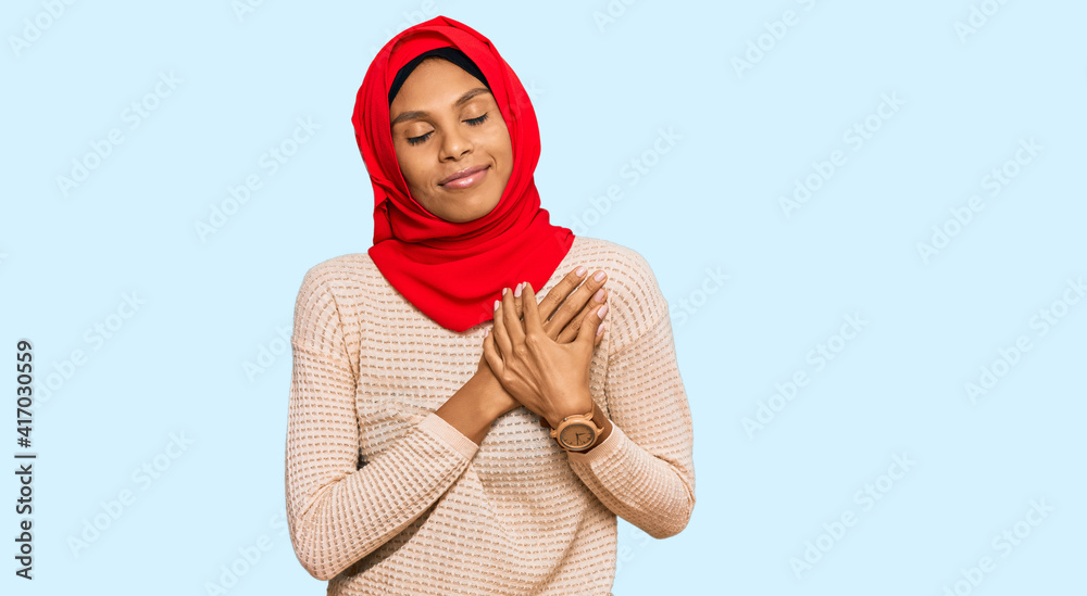 Young african american woman wearing traditional islamic hijab scarf smiling with hands on chest with closed eyes and grateful gesture on face. health concept. Young african american woman wearing traditional islamic hijab scarf smiling with hands on chest with closed eyes and grateful gesture on face. health concept.