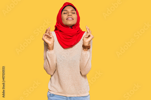Young african american woman wearing traditional islamic hijab scarf gesturing finger crossed smiling with hope and eyes closed. luck and superstitious concept.