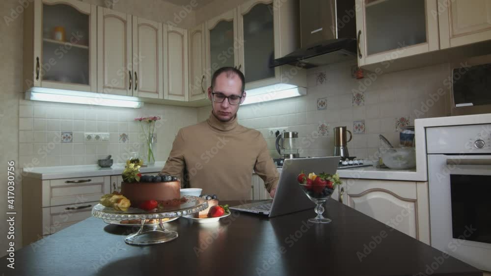 Man in glasses works on notebook and drinks tea at kitchen Man in glasses works on notebook and drinks tea at kitchen