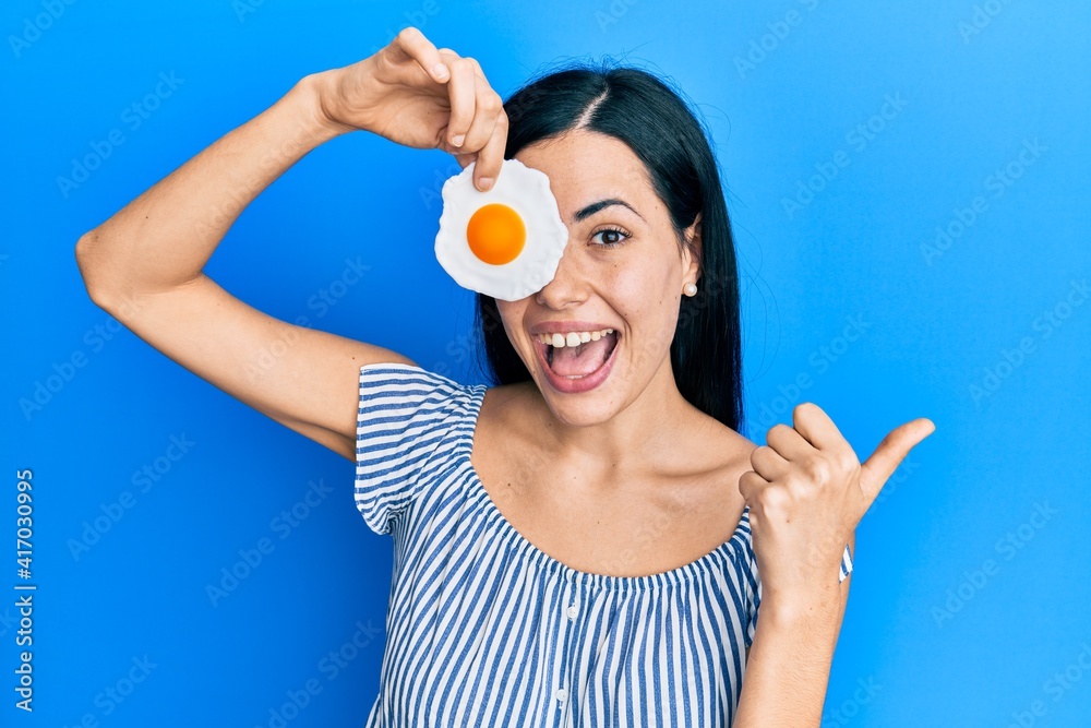 Beautiful young woman holding fried egg pointing thumb up to the side smiling happy with open mouth Beautiful young woman holding fried egg pointing thumb up to the side smiling happy with open mouth