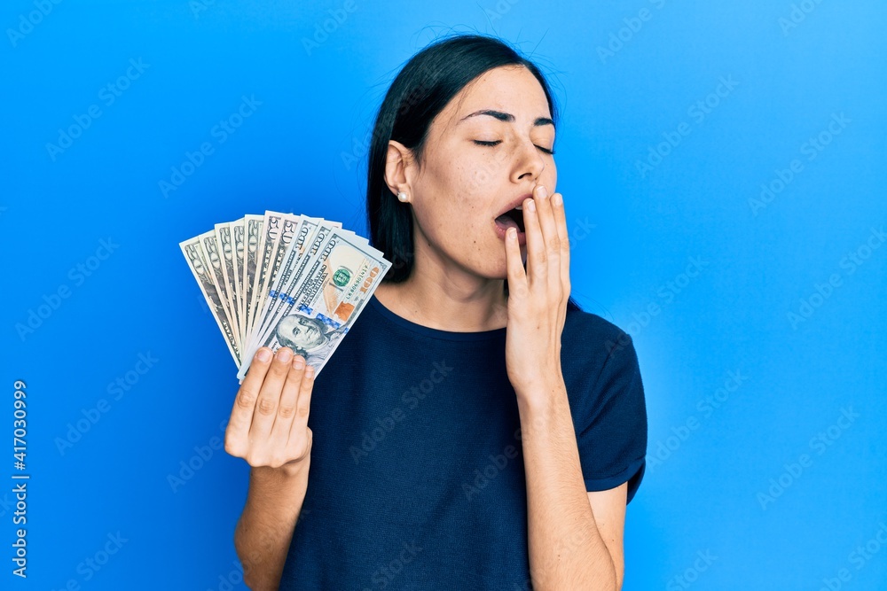 Beautiful young woman holding dollars bored yawning tired covering mouth with hand. restless and sleepiness. Beautiful young woman holding dollars bored yawning tired covering mouth with hand. restless and sleepiness.