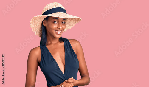 Young african american woman wearing swimsuit and summer hat with hands together and crossed fingers smiling relaxed and cheerful. success and optimistic