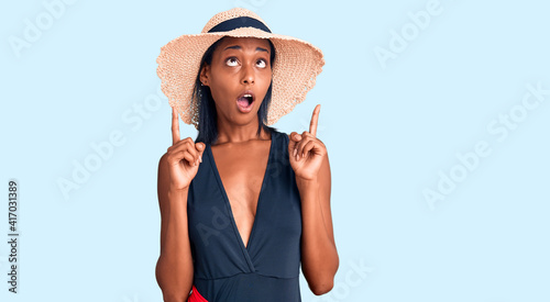 Young african american woman wearing swimsuit and summer hat amazed and surprised looking up and pointing with fingers and raised arms. photo