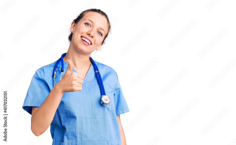 Young beautiful blonde woman wearing doctor uniform and stethoscope doing happy thumbs up gesture with hand. approving expression looking at the camera showing success. Young beautiful blonde woman wearing doctor uniform and stethoscope doing happy thumbs up gesture with hand. approving expression looking at the camera showing success.