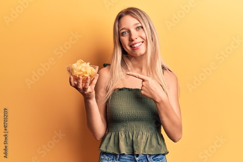 Young blonde woman holding potato chip smiling happy pointing with hand and finger