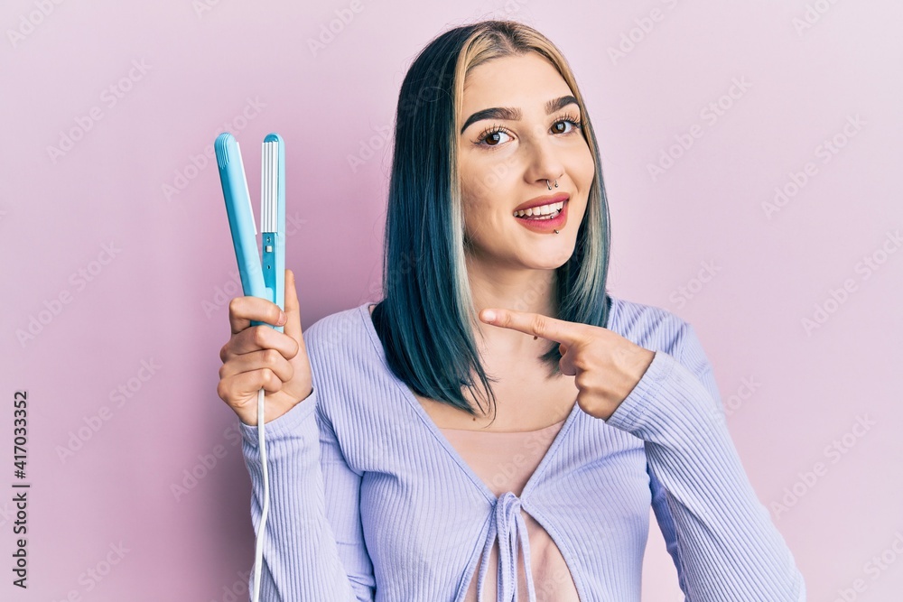 Young modern girl holding hair straightener smiling happy pointing with hand and finger Young modern girl holding hair straightener smiling happy pointing with hand and finger