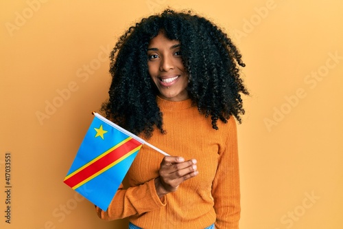African american woman with afro hair holding democratic republic of the congo flag looking positive and happy standing and smiling with a confident smile showing teeth